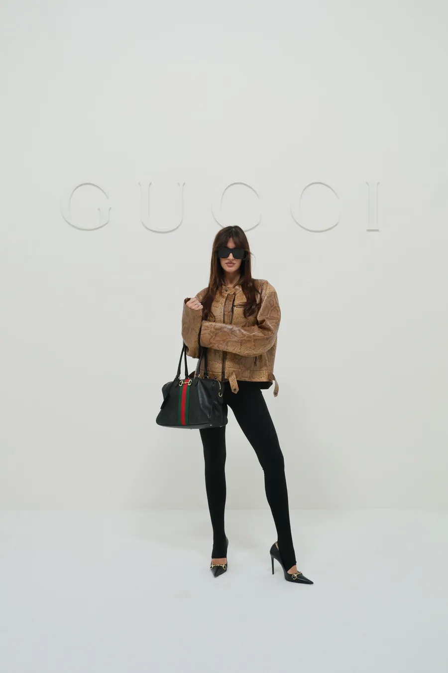 Gucci The Store Show: Frau in camelfarbener Jacke und schwarzer Hose mit schwarzer Gucci-Handtasche