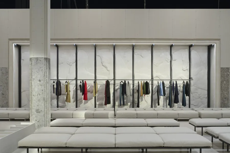 Gucci The Store Show: lange Kleiderstange vor einer weißen Reliefwand, davor Sitzbänke und eine Steinsäule